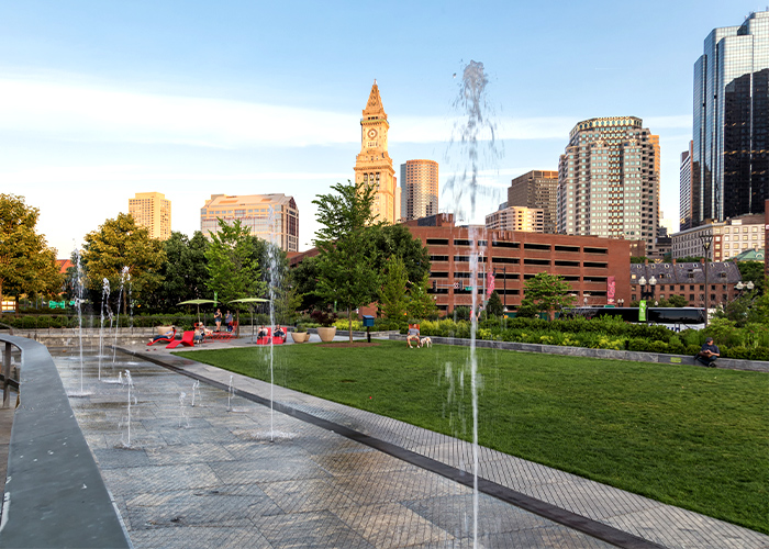 The Rose Fitzgerald Kennedy Greenway, a park with a stone pathway with fountains of water shooting up from the ground in Boston. Beside the path, there is a rectangular grass area with a person sitting in the corner on a red lawn chair, and behind it, there are chairs and umbrellas with a few people around. The horizon line is filled with a picturesque cityscape with tall buildings and structures.