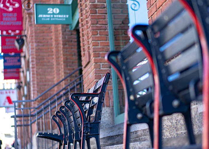 Black and red stadium seats along a brick wall at Fenway Park, baseball stadium in Boston. There are several red, blue, white and green banners hanging from the brick wall with text that is blurred.