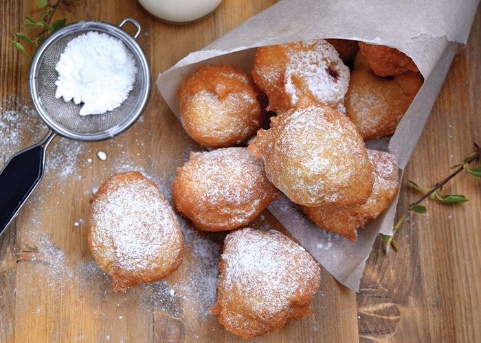 Several beignets, small, deep-fried dough with dustings of powdered sugar on top, wrapped in a sheet of parchment paper on a wooden surface. There is a small metal strainer with powdered sugar in it to the left of the beignets.