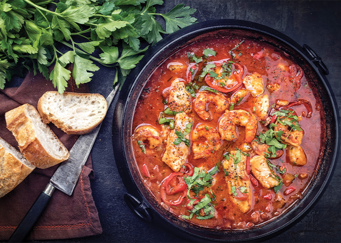 A dish containing Gumbo, a stew with okra, onions, celery, bell peppers, and shrimp mixed within a red sauce. To the left, there are a few slices of Italian bread, and a large clove of basil.