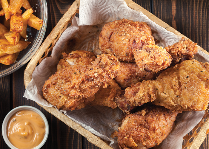 A basket filled with several pieces of fried chicken, with a small cup with a yellow dipping sauce and a bowl of french fries.