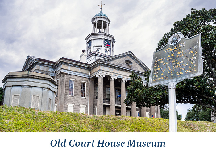 A ground-up view of the Old Courthouse Museum, a large building with two story columns by the entrance, and an open dome with a compass on the top in Vicksburg. At the bottom, there is blue text overlay on a white background that reads Old Court House Museum.