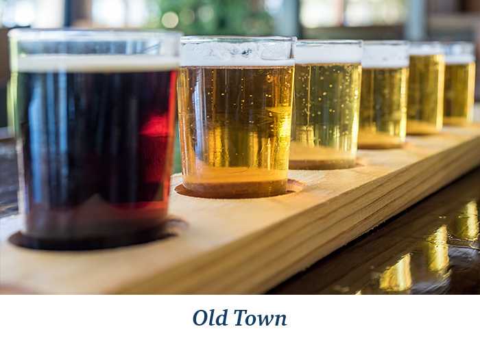 A line of craft beers in a wooden flight board at Cottonwood Public House in Old Town Vicksburg. One beer is a dark red color, while the other five are golden. At the bottom, there is blue text overlay on a white background that reads Old Town.