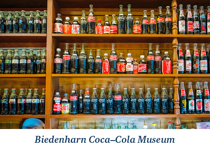 A wooden wall of shelves containing historic glass Coca-Cola bottles in a perfectly organized line at the Biedenharn Coca–Cola Museum in Vicksburg. At the bottom, there is blue text overlay on a white background that reads Biedenharn Coca–Cola Museum.