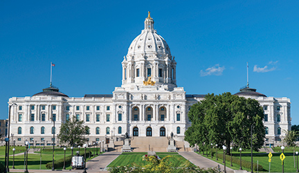 The Minnesota State Capitol Building in St. Paul. The large white building has a towering white marble dome in the center, and a flag pole on each end of the building. There are two concrete paths on each side of a perfectly maintained lawn, leading up to several stairs.