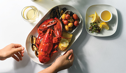 A plate with classic New England cuisine - a bright red lobster, two pieces of corn on the cob, and a small bowl of vegetables, and a small plate beside it with four lemon slices and a dipping bowl with melted butter, served on American Cruise Lines.