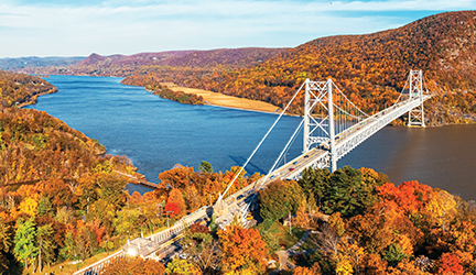 An aerial view of Bear Mountain Bridge across the Hudson River, and the hills covered in trees with vibrant red, orange, and yellow leaves on each side of the river during prime fall foliage season.