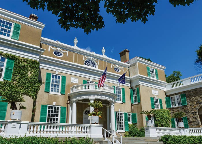 The Franklin D. Roosevelt Presidential Library and Museum, a large, tan building with bright green shutters, a half-circle balcony above the front door with an American flag flying from the railing, and vines and bushes thoughtfully placed around the entry way in Hyde Park.