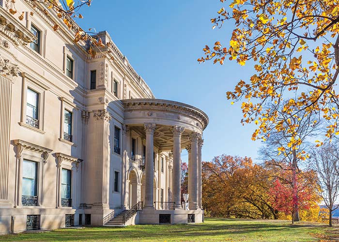 Vanderbilt Mansion National Historic Site in Hyde Park, a three-story, cream colored mansion with four tall pillars in front of the entryway stairs. There are several trees with orange and red leaves in the distance.