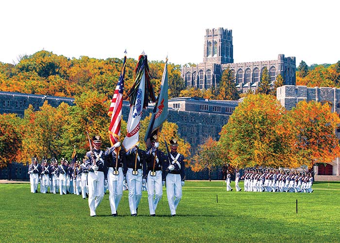 Five military soldiers are walking in uniform on the lawn of West Point, while the three in the middle are holding flags and the two on the outside are holding large guns. There are two groups of soldiers behind them and to the right, walking in the same formation. The United States Military Academy, a large, brick building is on a hill in the distance, surrounded by thick green trees.