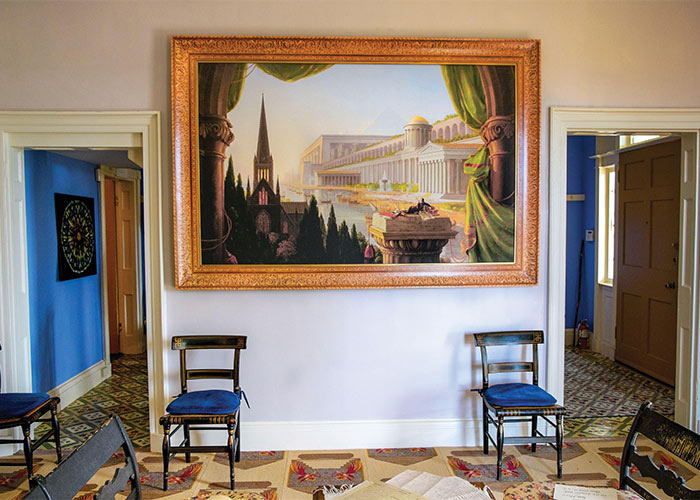 A large painting of a Hudson River region cityscape with a thick gold frame, painted by Thomas Cole on display at the Hudson River School Art Trail in Catskill. There are two blue chairs directly below the painting, and two open doors on each side leading to a separate room.