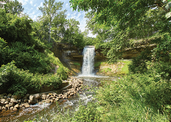 Minnehaha Falls, a small waterfall that leads to a stream lined with stones and thick greenery on each side in St. Paul.