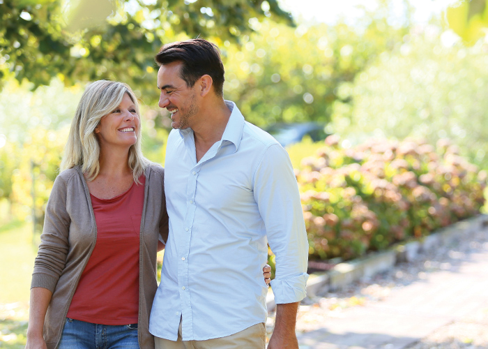A couple smiling at each other with their arms around one another while walking through Mounds Park. There are trees and flower bushes in the background.