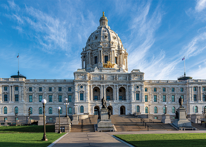 The Minnesota State Capitol Building in St. Paul. The large white building has a towering white marble dome in the center, and a flag pole on each end of the building. There are two large statues at the bottom of the many stairs.