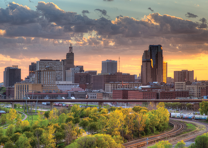 A landscape view of St. Paul, with a focus on Summit Avenue, where there are several large buildings and skyscrapers under an orange sunset. In the foreground, there are train tracks surrounded by several tall trees.