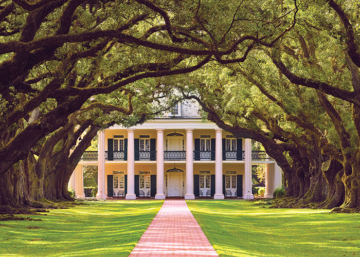 A historic plantation in Louisiana, Oak Alley Plantation, has two story pillars on all sides, grand porches, and is set amongst beautiful grounds and gardens with a red brick pathway with large oak trees on each side leading to the front door.