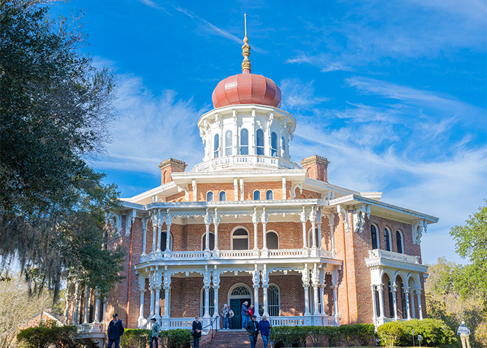 Longwood, a large, octagonal brick mansion in Natchez with a red byzantine onion-shaped dome with ornate white porches, surrounded by trees, under a blue sky. People are walking in the front yard.