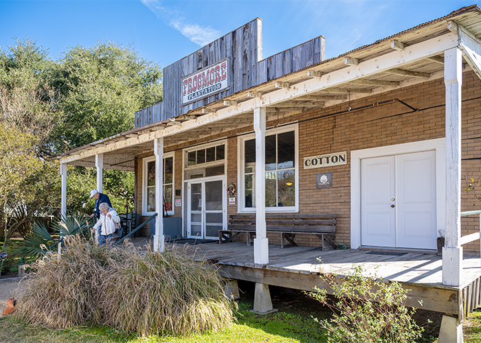 Frogmore Plantation, a historic brick building with two large windows, two glass doors, two white doors, a wooden porch, and a white sign that reads Cotton in Natchez. There are two American Cruise Lines guests walking down the stairs from the porch.