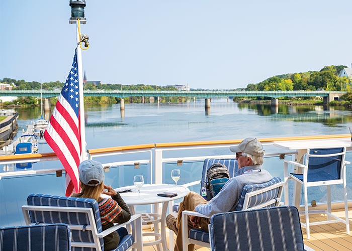 A couple sits at a table beside an American flag on an American Riverboat's sun deck looking out at the Mississippi River.
