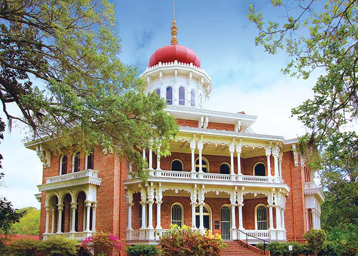 Longwood, a large, octagonal brick mansion in Natchez with a red byzantine onion-shaped dome with ornate white porches, surrounded by trees, under a blue sky. People are walking in the front yard.