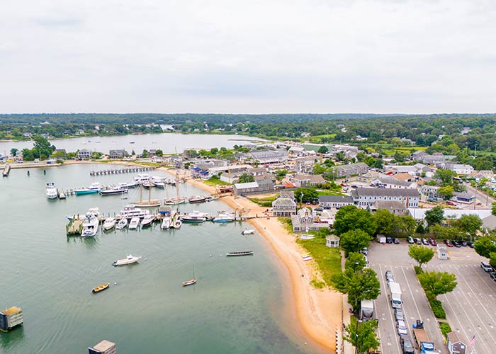 An aerial view of Vineyard Haven, a large port on Martha's Vineyard with several boats docked along the water. There is a small, sandy beach along the water with several buildings and trees stretching for miles in the distance.