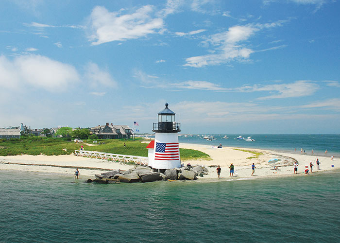 Brant Point Lighthouse, a small, white lighthouse on Nantucket Island with a large American flag on its side surrounded by rocks on the edge of the beach, along the water of Vineyard Sound. There is a ramp leading to the lighthouse from the grassy area behind the sand. People are fishing on the edge of the sand, and there are boats on the water and a large building in the middle of the island.