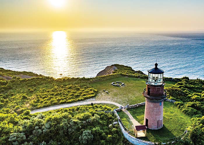 An aerial view of Gay Head Lighthouse, a small, red brick lighthouse on top of a hill in the center of a perfectly maintained grassland, where there are several trees and bushes surrounding the grass area in Martha's Vineyard. Beyond the hill is a breathtaking view of the New England coast with the sun reflecting on the water.