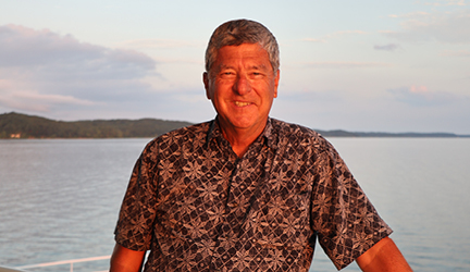 Jim Zim, an American Cruise Lines guest and social media influencer, smiling for a photo with the Tennessee River in the background. He is wearing a brown Hawaiian shirt with several white flowers covering it.