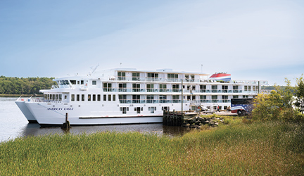 American Eagle, a small white Coastal Cat cruise ship docked in Bath. There is tall grass in the foreground, and an American Cruise Lines tour bus beside the small cruise ship.