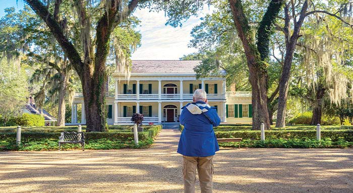 A man taking a photograph while standing outside of the historic St. Francisville landmark, the Rosedown, a yellow house with bright green shutters and two-story white pillars with a brick pathway surrounded by perfectly trimmed bushes and large trees leading to the red door.