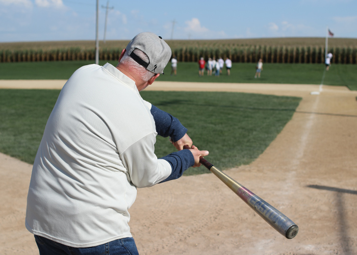 An older man swinging a baseball bat at the baseball field that was the set of the movie Field of Dreams, with faded white foul lines in the dirt, and trimmed grass in the center of the field and along the outfield. In the distance, a group of people are standing in right field.