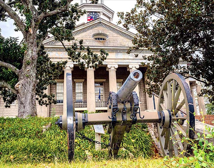 A cannon with two large wheels is pictured in front of the historic Old Warren County Courthouse, a light brick building with pillars out front in Vicksburg.