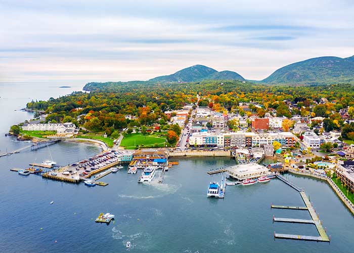 A small boat docked beside a long bridge on the Maine coast, with views of the autumn leaves on the full trees. There is a house on the shore that also has a long bridge leading out to the water.