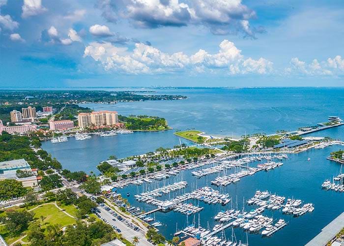 An aerial view of boats docked in a marina in St. Petersburg with the bright blue Gulf Coast waters spanning for miles in the distance. There are several buildings and trees on the shoreline.