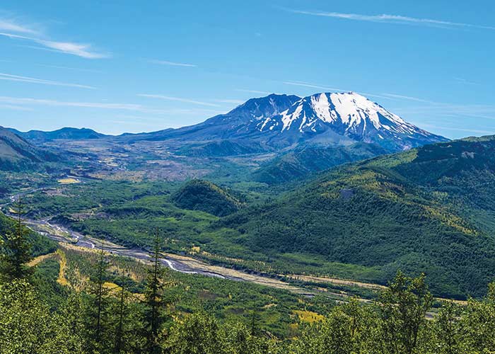 An aerial view of the snow-capped Mount St. Helens in Kalama, with small hills covered in grass and trees in the foreground and a small path running through the center.