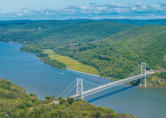An aerial view of a long bridge across the Hudson River, and the hills covered in trees on each side of the river.
