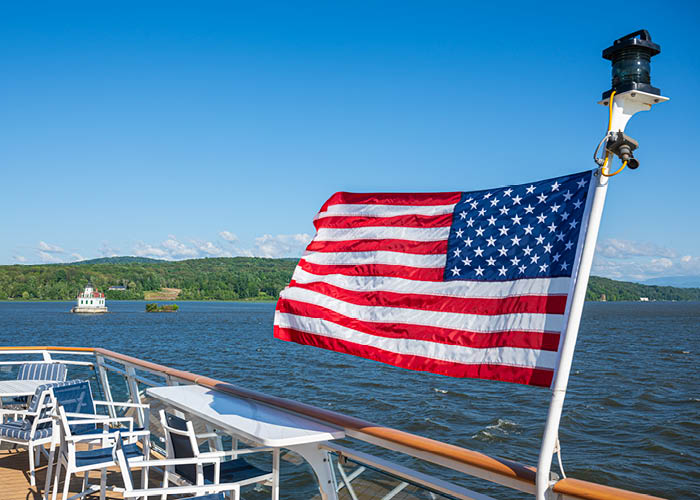 American flag on an American Cruise Lines ship, with chairs visible on the deck, and the river, green hills.