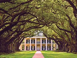 A historic plantation in Louisiana, Oak Alley Plantation, has two story pillars on all sides, grand porches, and is set amongst beautiful grounds and gardens with a red brick pathway with large oak trees on each side leading to the front door. 