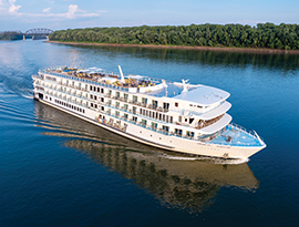 The American Serenade, an American Cruise Lines Riverboat gliding along the Mississippi River, with the tree-filled shore and a bridge in the background. 