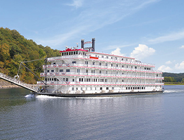 American Heritage paddlewheeler gliding along the Mississippi River. The white cruise ship is decorated with patriotic red white and blue bunting flags on the front of every railing.