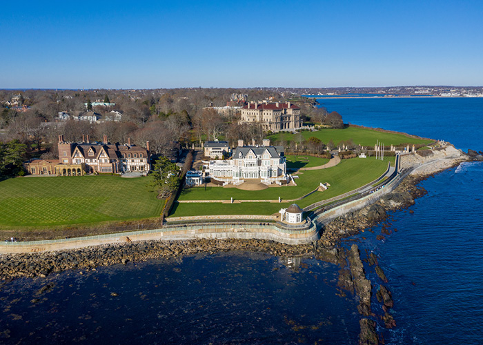 An aerial view of Anglesea, Breakers, and Fairholme Mansions in Newport. Anglesea mansion is tan with white shutters and a grey roof, and there is a gazebo at the edge of the large lawn, Fairholme mansion is brick with a dark brown roof, Breakers mansion appears as the tallest of the three, and is tan with a dark red roof. All three mansions have expansive front lawns that end at the waterline.