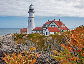 Portland Head Light, a white lighthouse next to a large white building with a red roof and green trim, on the edge of a rocky cliff beside Casco Bay. There are several people walking alongside the lighthouse.
