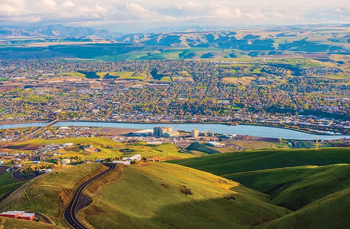 An aerial view of the city of Clarkston. In the foreground, there are grassy hills that lead up to the large cityscape in the center. In the far distance, there are several mountains spanning several miles.