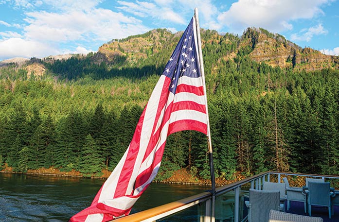 A vibrant American flag flying from the glass railing on the deck of an American Riverboat cruise ship while sailing along the Columbia River. Alongside the river, there is a cliff with thick forests spanning up the side.