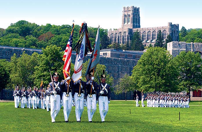 Five military soldiers are walking in uniform on the lawn of West Point, while the three in the middle are holding flags and the two on the outside are holding large guns. There are two groups of soldiers behind them and to the right, walking in the same formation. The United States Military Academy, a large, brick building is on a hill in the distance, surrounded by thick green trees.