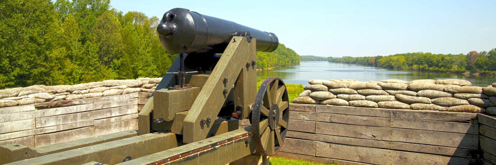A large black cannon in Fort Donelson National Battlefield that sits on a wooden stand is pointed directly towards the Mississippi River. The cannon is surrounded by a short wooden fence with sand bags on top of it.