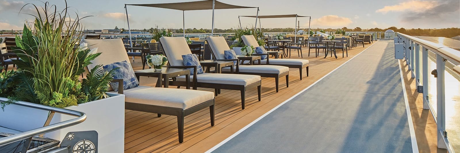 Several white lounge chairs with blue patterned pillows are placed next to a large plant in a grey, square pot along the sky walk on the sun deck of an American Riverboat.