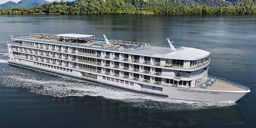 American Anthem, an American Cruise Lines American Riverboat small ship, cruising along the Columbia River, where the shore with small hills and trees is in the near distance.