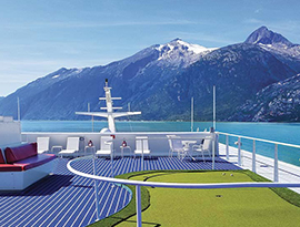 The sun deck of a American Constellation, an American Cruise Lines small cruise ship sailing on the crystal blue waters of the Wrangell Narrows with snow-capped mountains in the near distance. The sun deck has a perfectly maintained putting green, red benches, white lounge chairs, and a small round table surrounded by chairs.