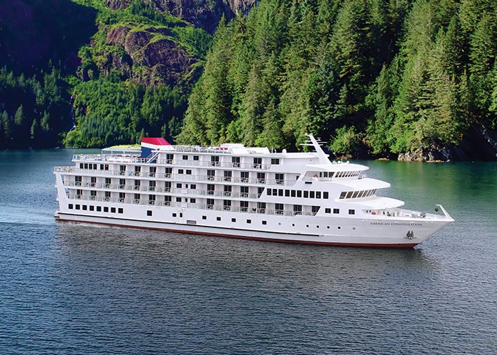 American Constellation, an American Cruise Lines small cruise ship sailing along the Alaska coast. In the near distance, there are dense, tree-filled forests along the side of a steep cliff.